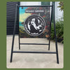 A black metal A-frame sidewalk sign displaying a coffee-themed graphic with the text 'PROUDLY SERVING' and a circular logo reading 'mahalia coffee', photographed outdoors on a concrete surface with plants and a building wall in the background.