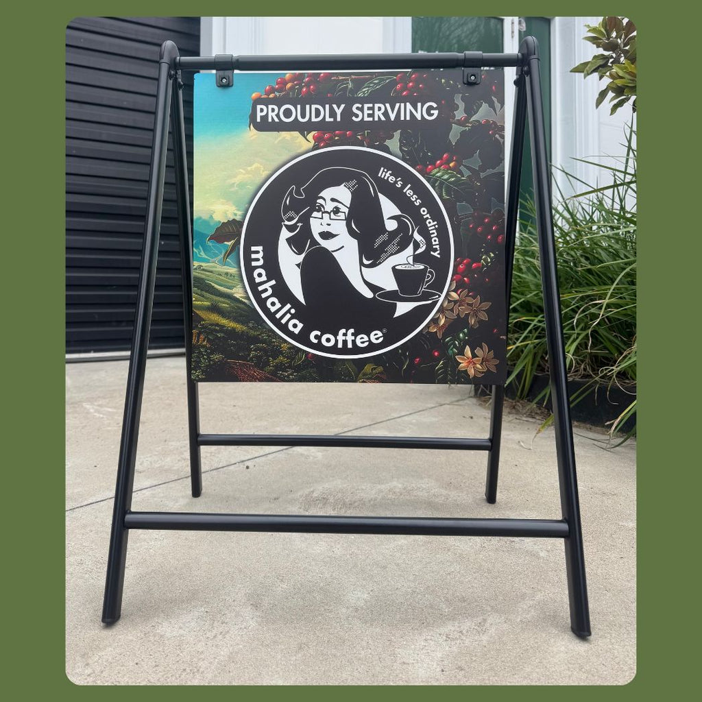 A black metal A-frame sidewalk sign displaying a coffee-themed graphic with the text 'PROUDLY SERVING' and a circular logo reading 'mahalia coffee', photographed outdoors on a concrete surface with plants and a building wall in the background.