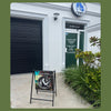 A storefront exterior with a black roller shutter on the left, a green door with signage, a round hanging logo sign, and a freestanding A-frame sidewalk sign in front. Plants line the right side of the entrance.