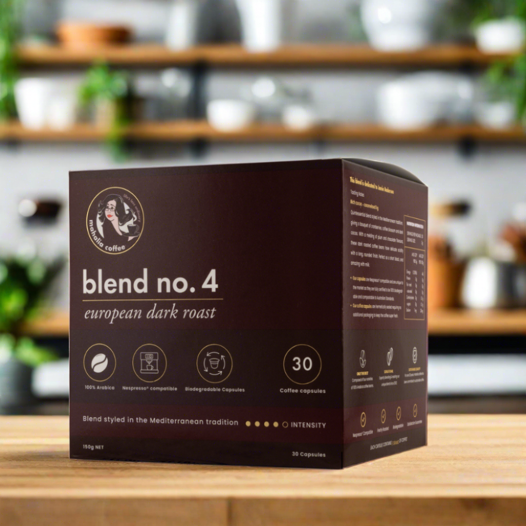 A box of Blend 4 coffee capsules, compatible with Nespresso machines, displayed on a wooden surface with a blurred background of indoor plants and wooden furniture.