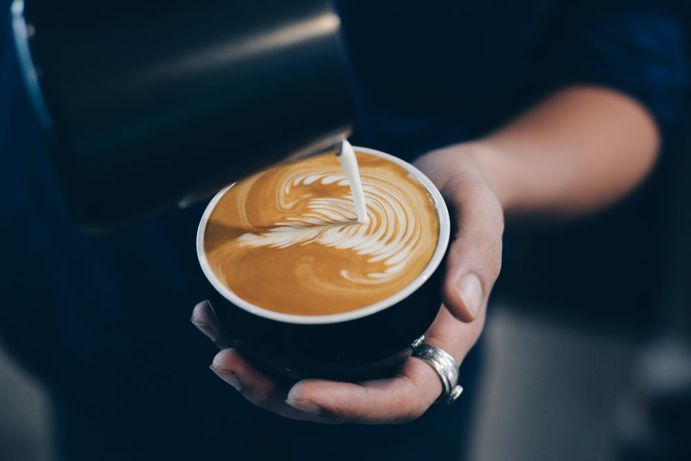 A close-up of a person's hand holding a paper cup of latte art while pouring milk from a metal pitcher into the coffee. The latte features a clean, swirling white foam pattern on a rich brown coffee base.